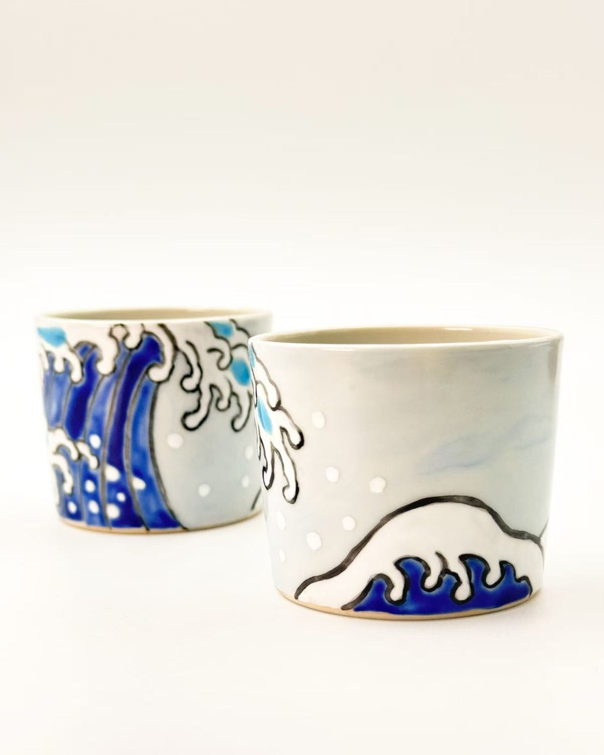 Two ceramic cups decorated with a blue and white wave pattern, reminiscent of a famous ocean wave painting, on a clean white background.